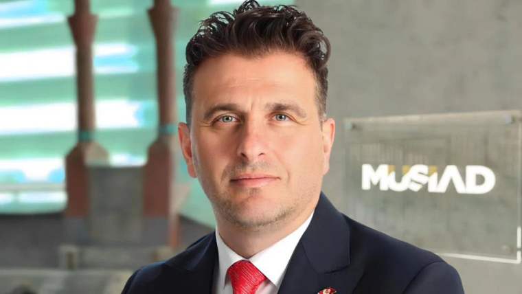 Kırmızı kravat takan bir adam, kameraya bakarak poz veriyor. Arka planda MÜSİAD logosu görünmekte.