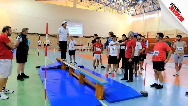 Bir grup genç, spor salonunda yetenek sınavına katılıyor; bir kişi denge tahtasında duruyor.