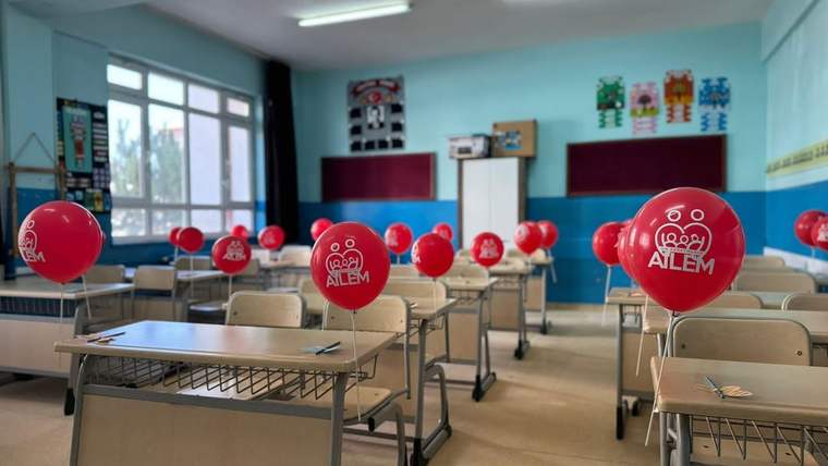Sınıfta sıraların arasına yerleştirilmiş, üzerinde “AİLEM” yazan çok sayıda kırmızı balon görülüyor.