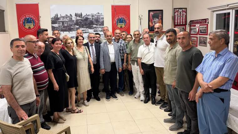 Bir grup insan, bir odada bir araya gelerek fotoğraf çektiriyor. Arka planda Sivas'a ait bir fotoğraf ve bayraklar var.