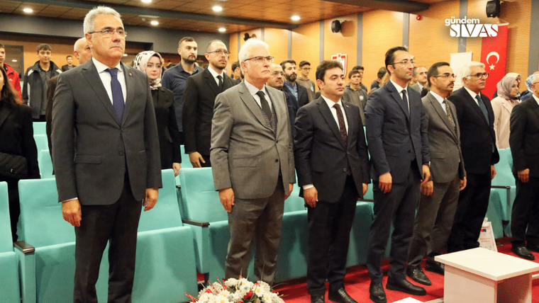 Bir etkinlikte, ayakta duran altı kişi ve arka planda gençlerden oluşan bir kalabalık yer alıyor.