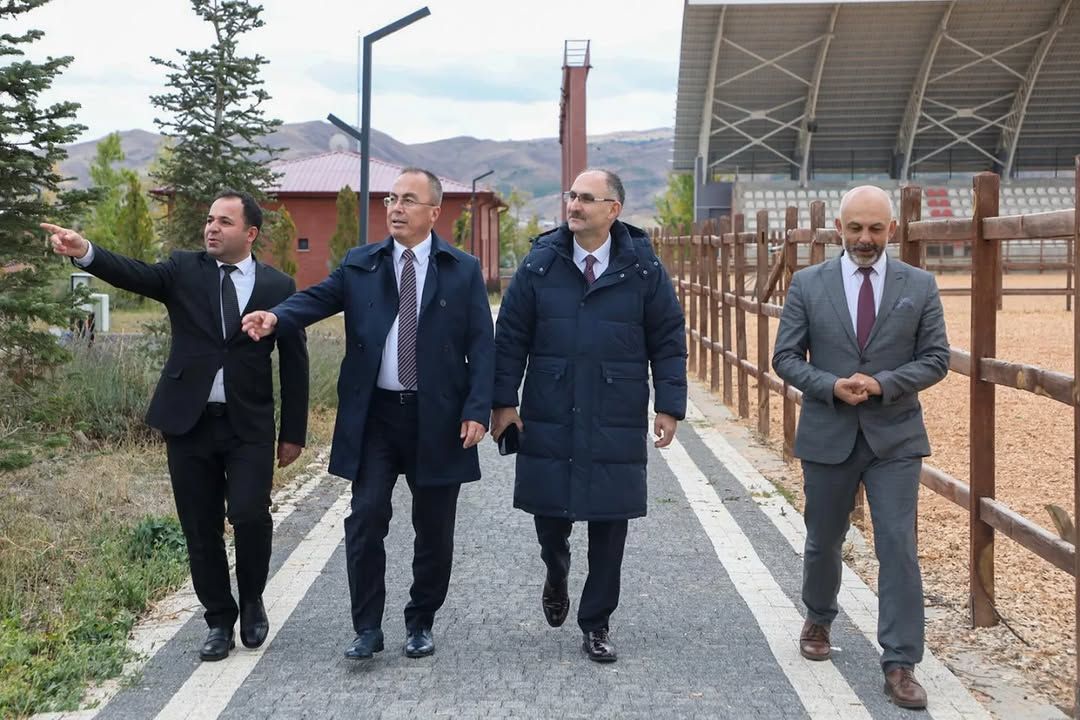 Taş döşeli yolda yürüyen dört erkekten biri ileriyi işaret ediyor; çevrede çit, ağaçlar ve dağlar görülüyor.