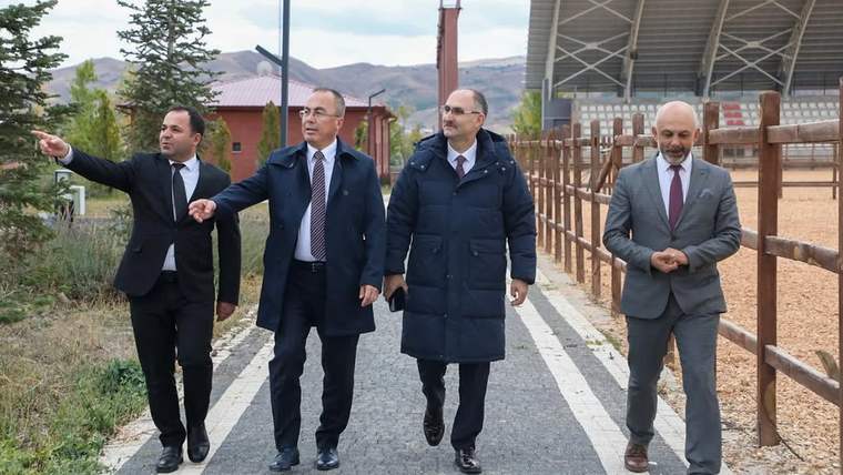 Taş döşeli yolda yürüyen dört erkekten biri ileriyi işaret ediyor; çevrede çit, ağaçlar ve dağlar görülüyor.