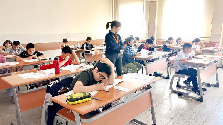 Sınıfta, masalarda oturan öğrenciler sınav yapıyor; öğretmen bir öğrencinin yanındadır.