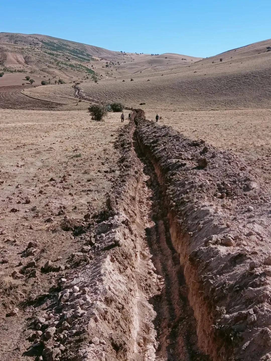Kurak bir tarlada uzanan derin hendek ve ileride yürüyen birkaç kişi görülüyor.