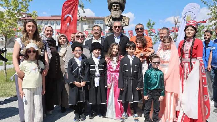 Büstün önünde, Türk bayrağı ve yöresel kıyafetler taşıyan çocuklarla yetişkinlerden oluşan grup duruyor.