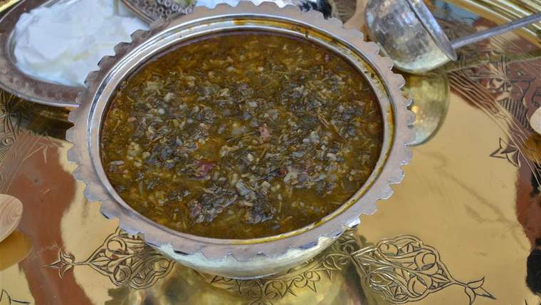 Süslemeli metal servis tepsisinde, doğranmış yeşilliklerle hazırlanmış sulu bir yemek ve yanında servis kapları bulunuyor.