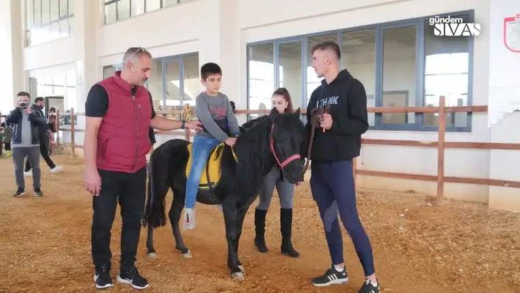 Bir çocuk, küçük bir atın üzerinde oturuyor; yanında iki yetişkin var.