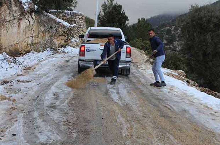 Karl manzaralı dağ yolunda iki kişi, karla kaplı zemine kürekle kum döken bir kamyonetin yanında çalışıyor.