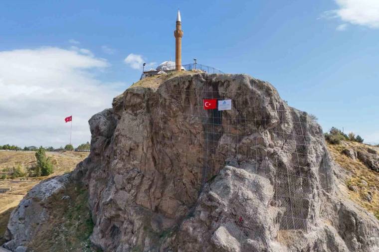 Kayalık tepenin üzerinde bir minare, kaya yüzeyinde Türk bayrağı ve metal ağ görülüyor.