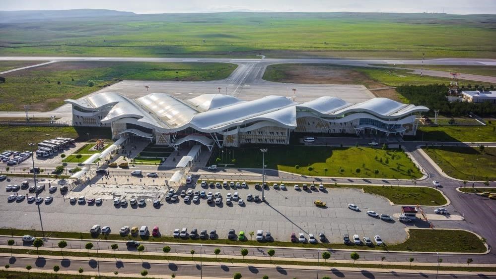 Havadan görünümde dalgalı çatılı havalimanı terminali, otoparkı ve arkasındaki pist bağlantıları görülüyor.