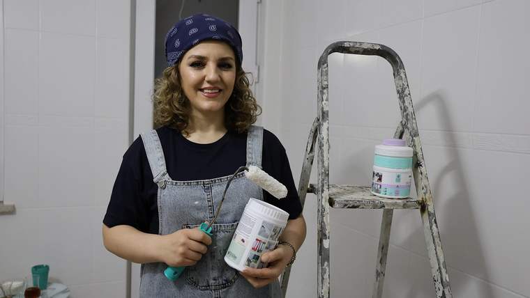 Başında bandana bulunan kadın, elinde boya rulosu ve boya kutusuyla merdivenin yanında duruyor.