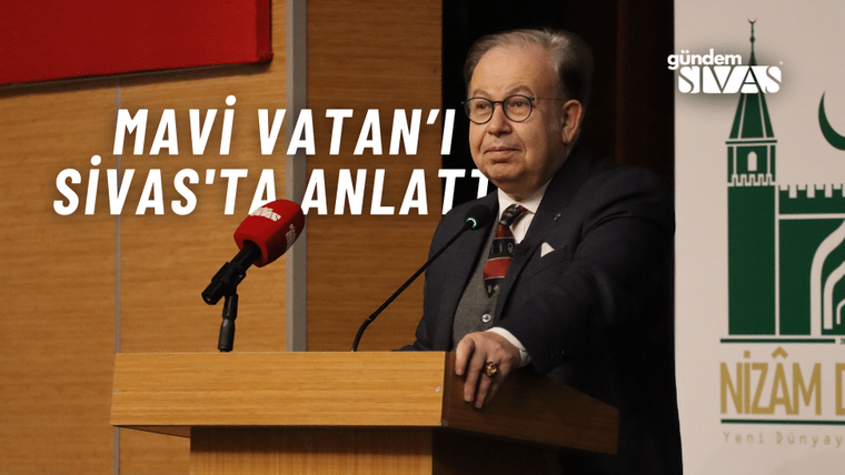 Gözlüklü bir kişi, ahşap kürsüde iki mikrofonun önünde duruyor; arkada yeşil amblemli pano var.