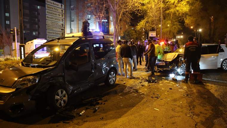 Gece aydınlatılmış caddede siyah bir otomobil, yanındaki araçlar, insanlar ve yanıp sönen ışıklar görülüyor.
