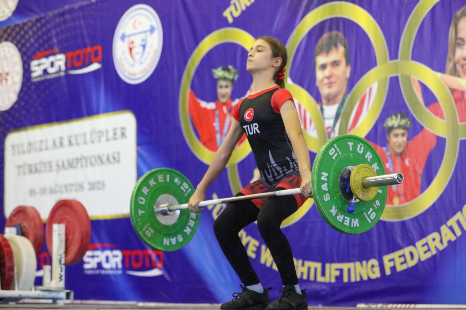 Halter kaldıran bir genç sporcu, arka planda halter federasyonu logosu ve olimpiyat halkaları var.