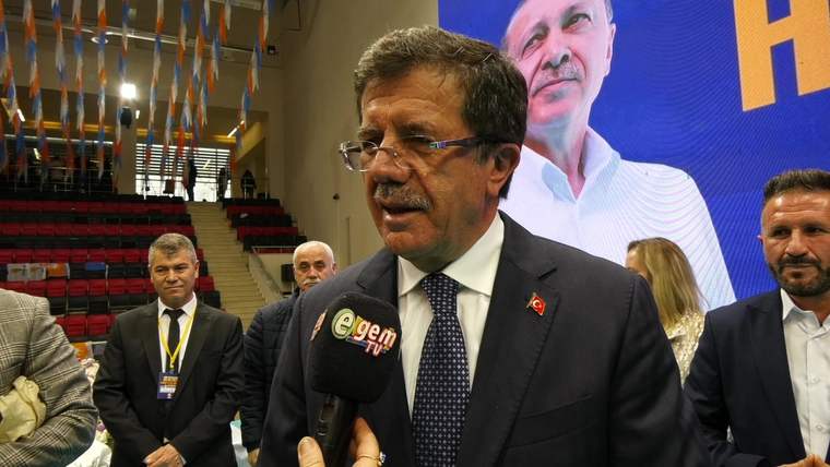 Mikrofonla konuşan bir kişi, arka planda büyük bir afiş yer alıyor.