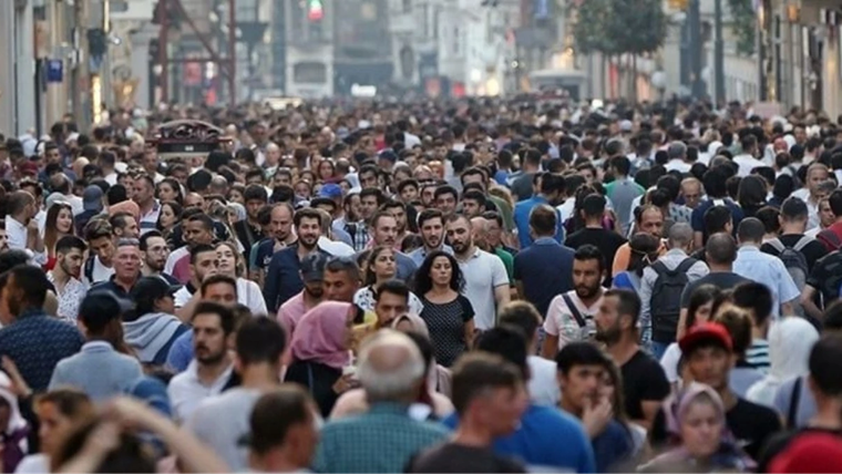 Geniş bir caddede çok sayıda insan kalabalık halinde yürürken, arka planda mağaza ve trafik ışıkları görülüyor.