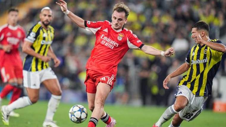 Kırmızı formalı futbolcu topa doğru ilerlerken, iki sarı-lacivert çubuklu oyuncu yanında mücadele ediyor.