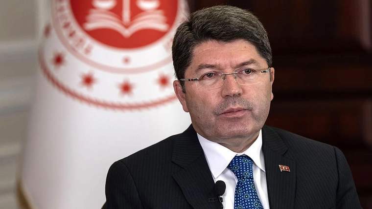 Gözlüklü bir kişi, takım elbise ve kravatla, yakasında Türk bayrağı rozeti ve önünde mikrofonla görülüyor.