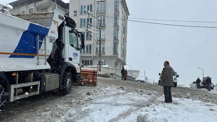 Kar yağışı altındaki yolda, üzerinde “CANİK” yazan bir kamyon ve turuncu iş makinesi görülüyor.