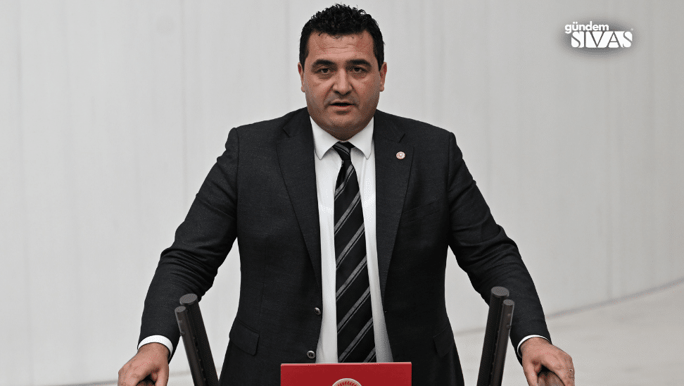 Kürsüde duran takım elbiseli bir kişi, önündeki kırmızı kapaklı kürsüyle konuşma yapıyor.