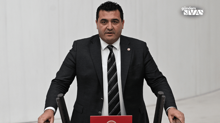 Kürsüde duran takım elbiseli bir kişi, önündeki kırmızı kapaklı kürsüyle konuşma yapıyor.