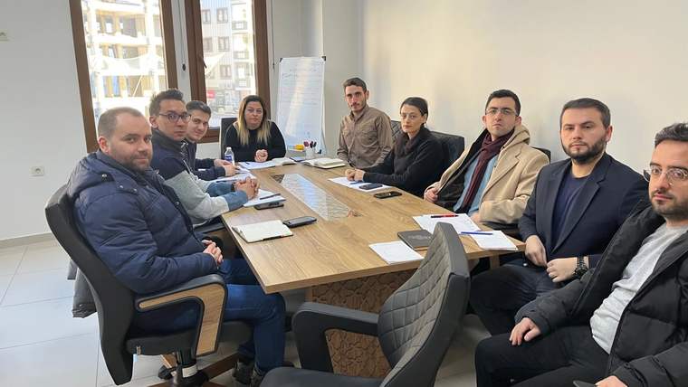 Toplantı masası çevresinde oturan sekiz kişi, önlerindeki belgeler ve bilgisayarlarla birlikte görülüyor.