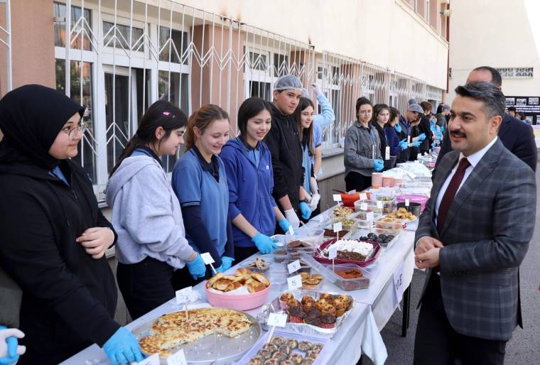 Okul önünde eldivenli öğrenciler, çeşitli yiyeceklerin bulunduğu masada stant başında duruyor; bir kişi yanlarında bekliyor.