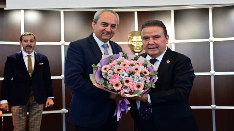 Çiçek buketi tutan bir kişi, diğerine gülümsüyor; arka planda bir heykel ve başka bir kişi görünüyor.