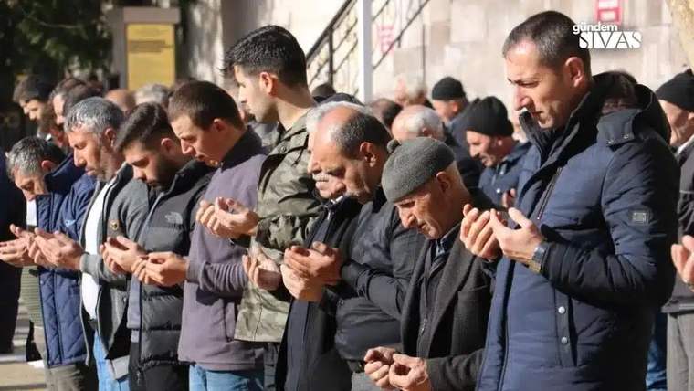 Bir grup erkek, ellerini açarak açık havada dua ediyor; arkalarında kalabalık ve bina cephesi görülüyor.