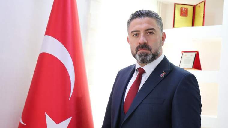 Kırmızı beyaz Türk bayrağının önünde, takım elbise giymiş bir adam duruyor.