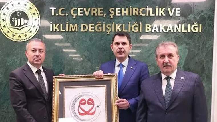 T.C. Çevre, Şehircilik ve İklim Değişikliği Bakanlığı tabelasının önünde üç kişi, bir çerçeve tutuyor.