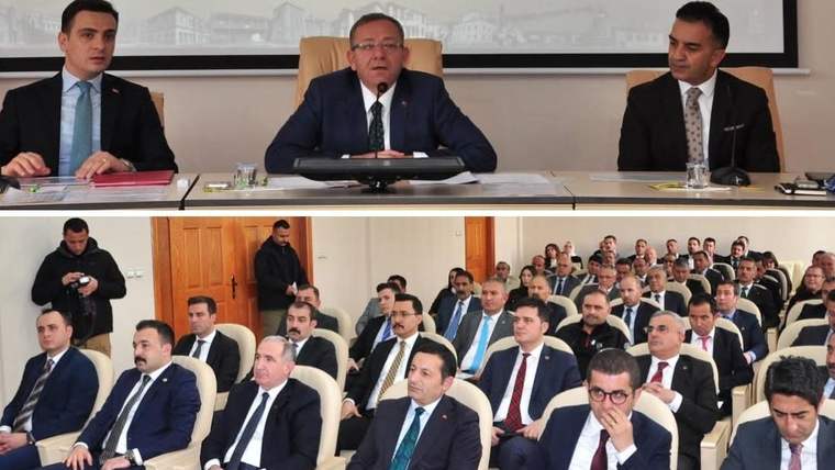 Toplantı salonunda masada oturan üç kişi ve karşılarında sıralar halinde oturan kalabalık bir grup görülüyor.