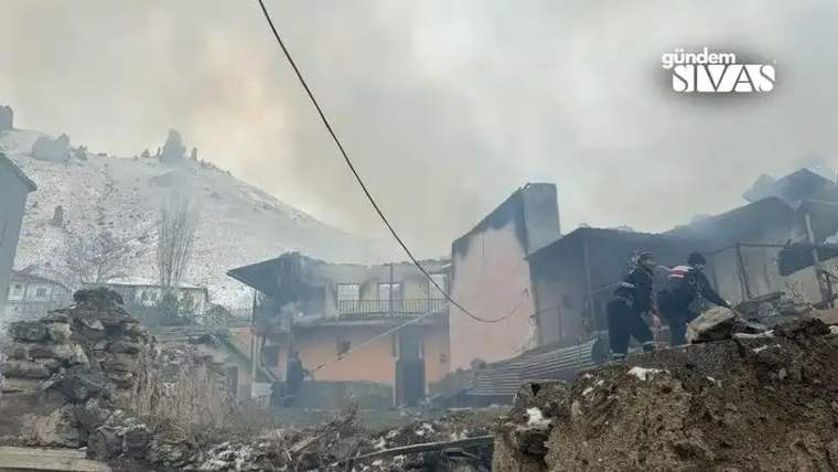 Yanmış evlerin kalıntıları arasında itfaiye ekipleri çalışıyor, arka planda dağlık alan görünüyor.