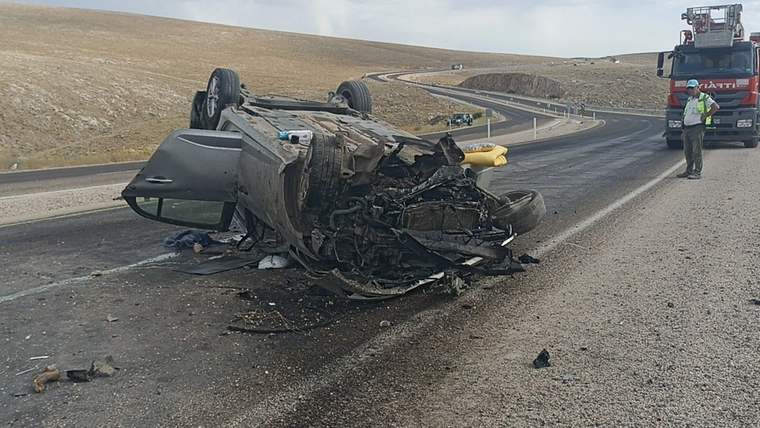 Bölünmüş yolda ters dönmüş, ağır hasar görmüş bir otomobilin çevresinde parçalar ve yol kenarında araçlar görülüyor.