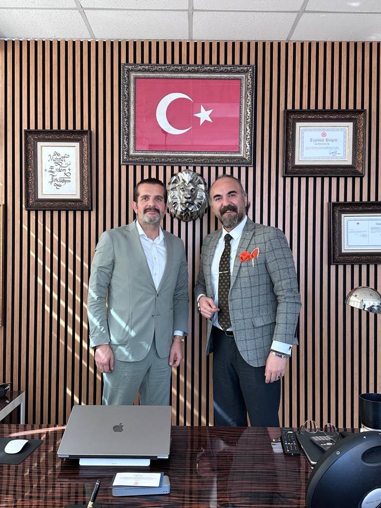 Ofis ortamında iki erkek, Türk bayrağı ve çerçeveli belgelerin önünde yan yana duruyor.