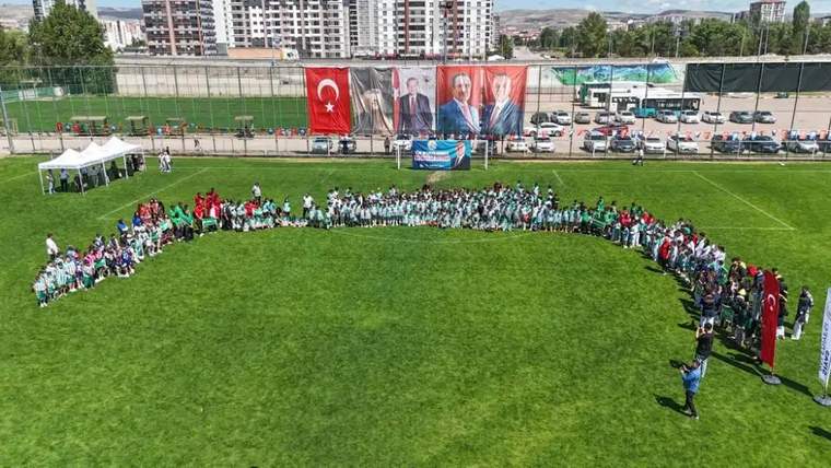 Yeşil bir alanda, spor okullarına katılan çocuklar ve eğitmenler bir araya gelerek grup oluşturmuş.