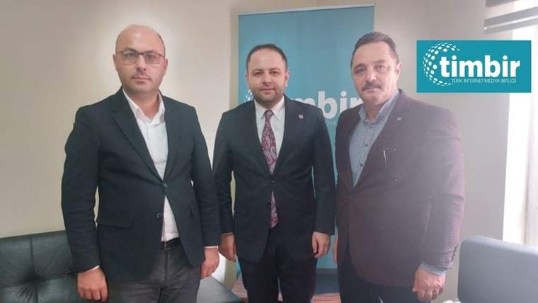 TİMBİR ofisinde, üç kişi bir arada duruyor; biri resmi kıyafet giymiş, diğerleri ise ceketli.