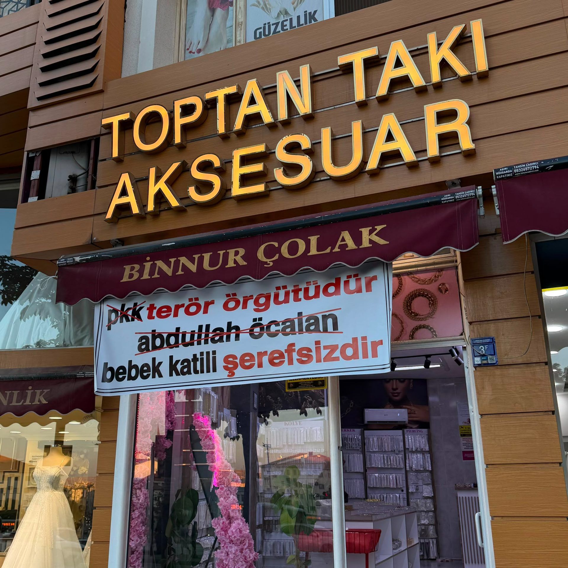 Takı ve aksesuar dükkânının cephesinde “Binnur Çolak” yazılı tente ve sloganlı bir pankart görülüyor.