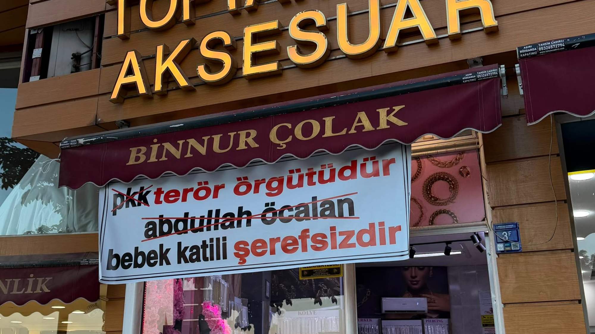 Takı ve aksesuar dükkânının cephesinde “Binnur Çolak” yazılı tente ve sloganlı bir pankart görülüyor.