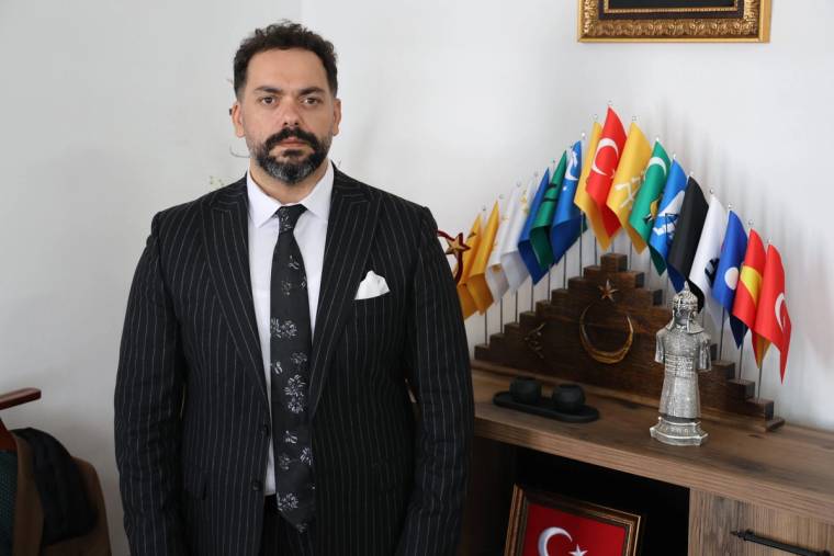 Kravat takmış bir adam, arkasında çeşitli bayrakların yer aldığı bir masa önünde duruyor.