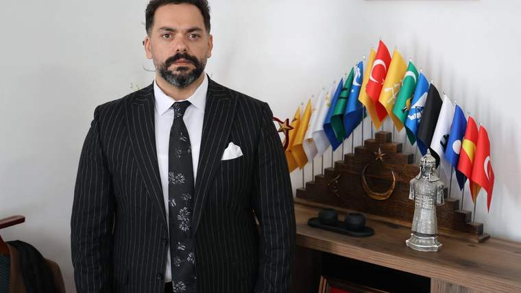 Kravat takmış bir adam, arkasında çeşitli bayrakların yer aldığı bir masa önünde duruyor.