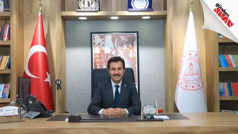 Türk bayrağı ve kırmızı amblemli beyaz bayrak önünde, çalışma masasının arkasında oturan bir kişi