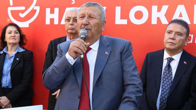Mikrofonla konuşan bir erkek, kırmızı yazılı pano önünde üç kişiyle birlikte duruyor.