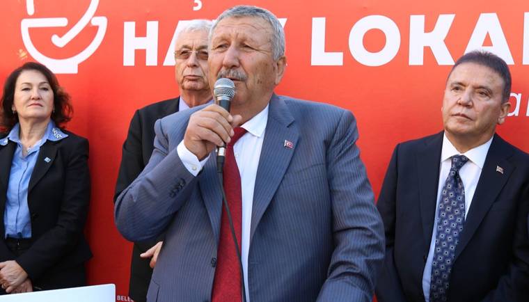 Mikrofonla konuşan bir erkek, kırmızı yazılı pano önünde üç kişiyle birlikte duruyor.