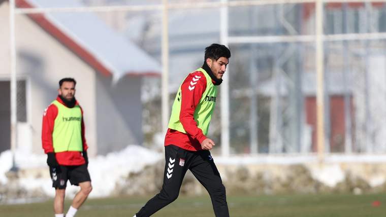 Yeşil yelekli iki futbolcu, karlı saha kenarında topla antrenman yaparken öndeki oyuncu top sürüyor.