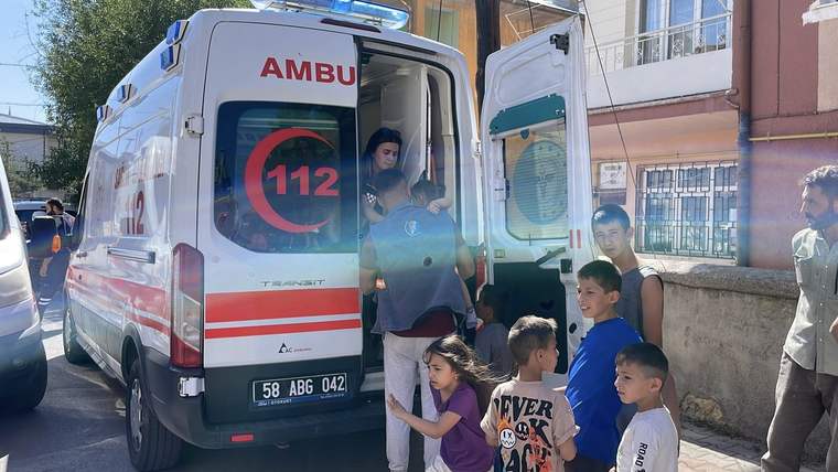 Açık arka kapısında bir kişinin bulunduğu ambulansın yanında, yetişkinler ve çocuklar kaldırımda duruyor.