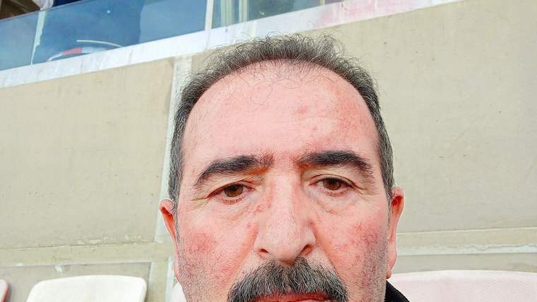 Bıyıklı, kısa saçlı bir adamın yüzü yakın planda görülüyor; arka planda camlı bir bölüm bulunuyor.