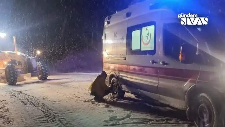 Karla kaplı yolda bir kişi, ambulansın lastiğini kontrol ediyor.