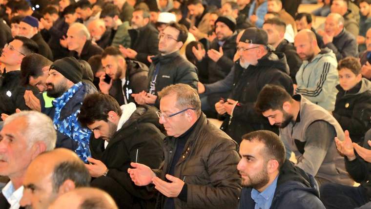 Kapalı bir alanda yan yana oturan kalabalık erkek topluluğu, ellerini açarak dua ediyor.